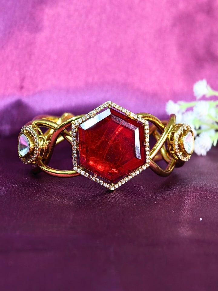 The Tahira Red Bracelet (Adjustable) features a large hexagonal Red stone at the center, with an american diamond border and two round kundan stones on each side, displayed on Purple satin fabric with white flowers in the background.