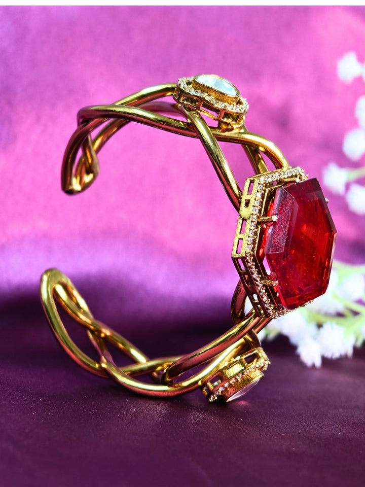 The Tahira Red Bracelet (Adjustable) features a large hexagonal Red stone at the center, with an american diamond border and two round kundan stones on each side, displayed on Purple satin fabric with white flowers in the background.