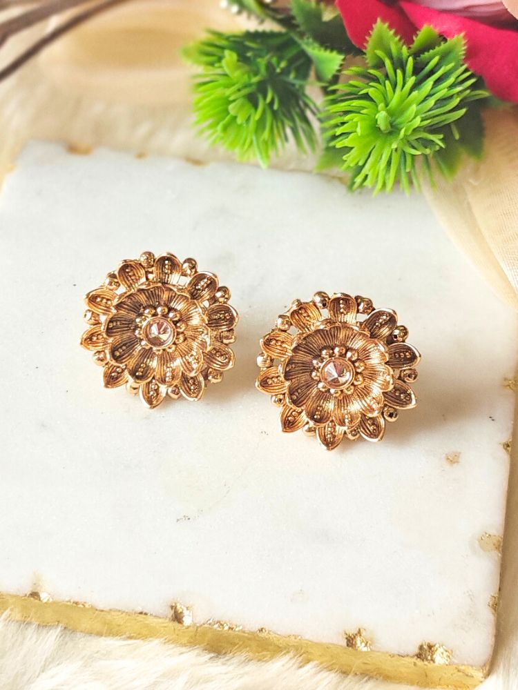 The Tanisha Gold stud Earrings, crafted from brass with gold floral designs featuring intricate petals and a central gemstone, are displayed on a white marble slab with blurred green leaves and a red flower in the background.