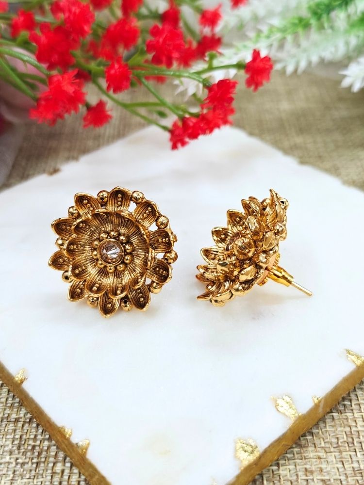 The Tanisha Gold stud Earrings, crafted from brass with gold floral designs featuring intricate petals and a central gemstone, are displayed on a white marble slab with blurred green leaves and a red flower in the background.