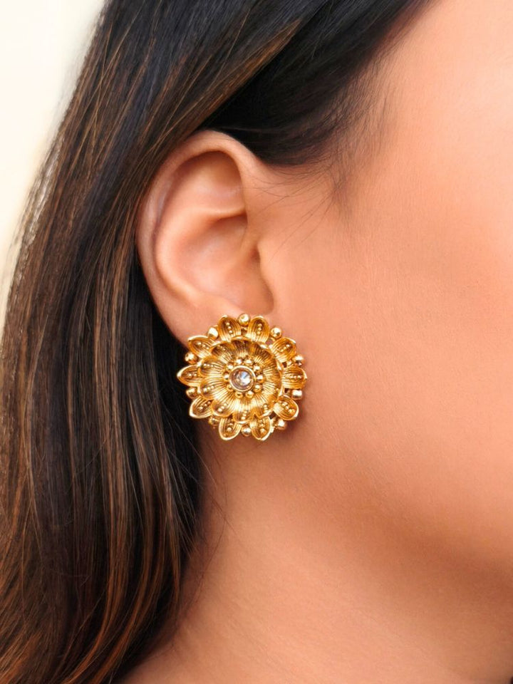  A women wearing The Tanisha Gold stud Earrings, crafted from brass with gold floral designs featuring intricate petals and a central gemstone. Indo-western dress styled with lightweight artificial American Diamond Studs earrings. Festive Diwali look styled with premium artificial American Diamond Studs earrings.