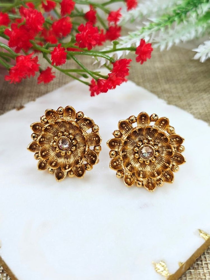 The Tanisha Gold stud Earrings, crafted from brass with gold floral designs featuring intricate petals and a central gemstone, are displayed on a white marble slab with blurred green leaves and a red flower in the background.