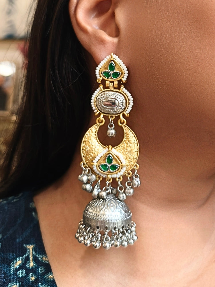 A close-up of a womans ear wearing Tapasya Green Earrings, ornate gold and silver with floral designs, colored stones, dangling silver beads, and a large intricate jhumka dome at the bottom. Long Oxidized earrings, german silver earrings, navratri earrings.  Jhumka earrings. Jhumki Earrings. 