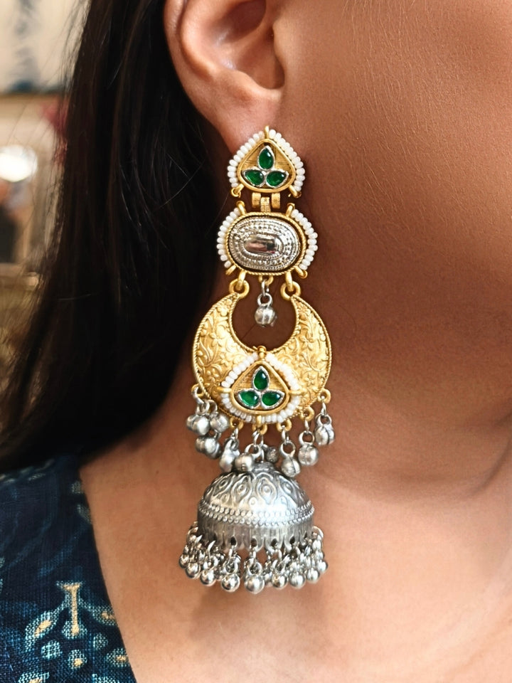 A close-up of a womans ear wearing Tapasya Green Earrings, ornate gold and silver with floral designs, colored stones, dangling silver beads, and a large intricate jhumka dome at the bottom. Long Oxidized earrings, german silver earrings, navratri earrings.  Jhumka earrings. Jhumki Earrings. 