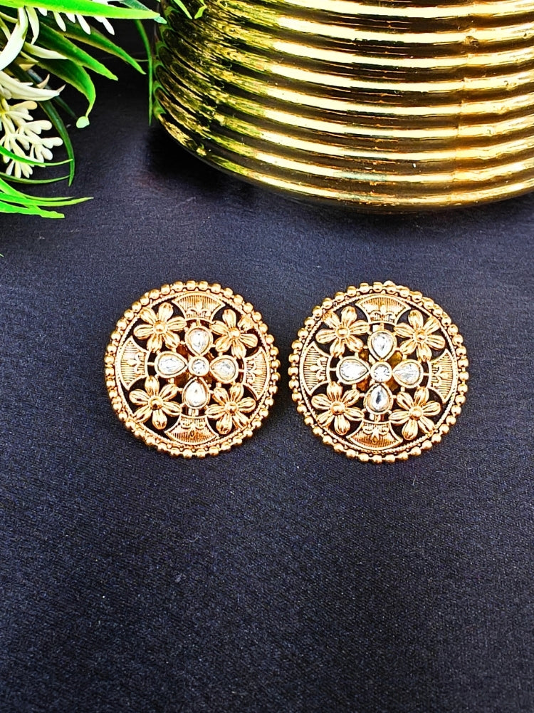 The Tarang Earrings are round, gold-toned studs featuring intricate floral designs and embedded American Diamond stones, showcased on a black fabric background near a gold ribbed container and green leaves.