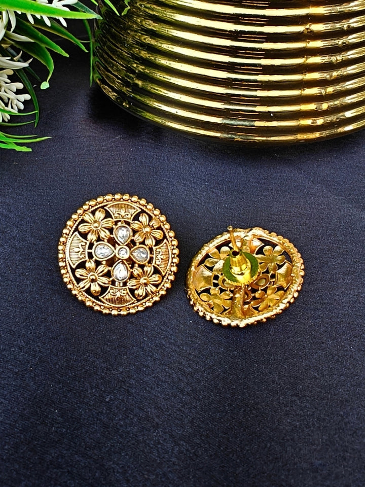 The Tarang Earrings are round, gold-toned studs featuring intricate floral designs and embedded American Diamond stones, showcased on a black fabric background near a gold ribbed container and green leaves.
