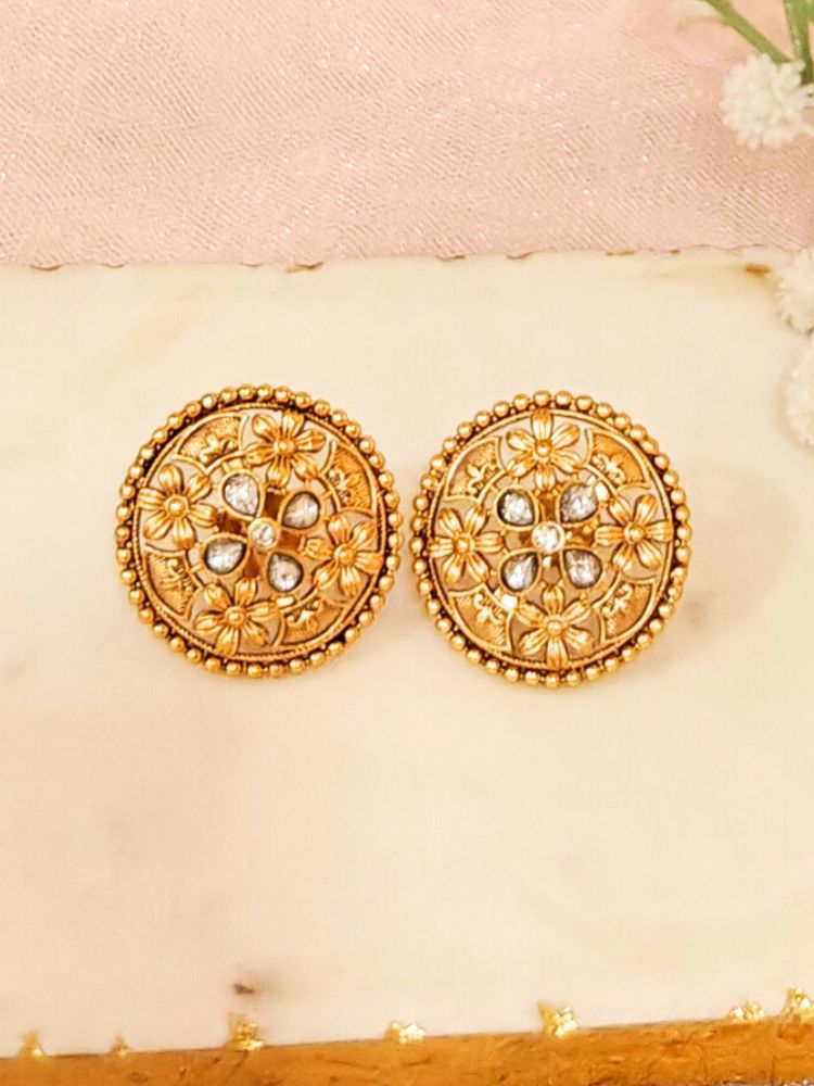 The Tarang Earrings are round, gold-toned studs featuring intricate floral designs and embedded American Diamond stones, showcased on a White Marble background with White Flower.