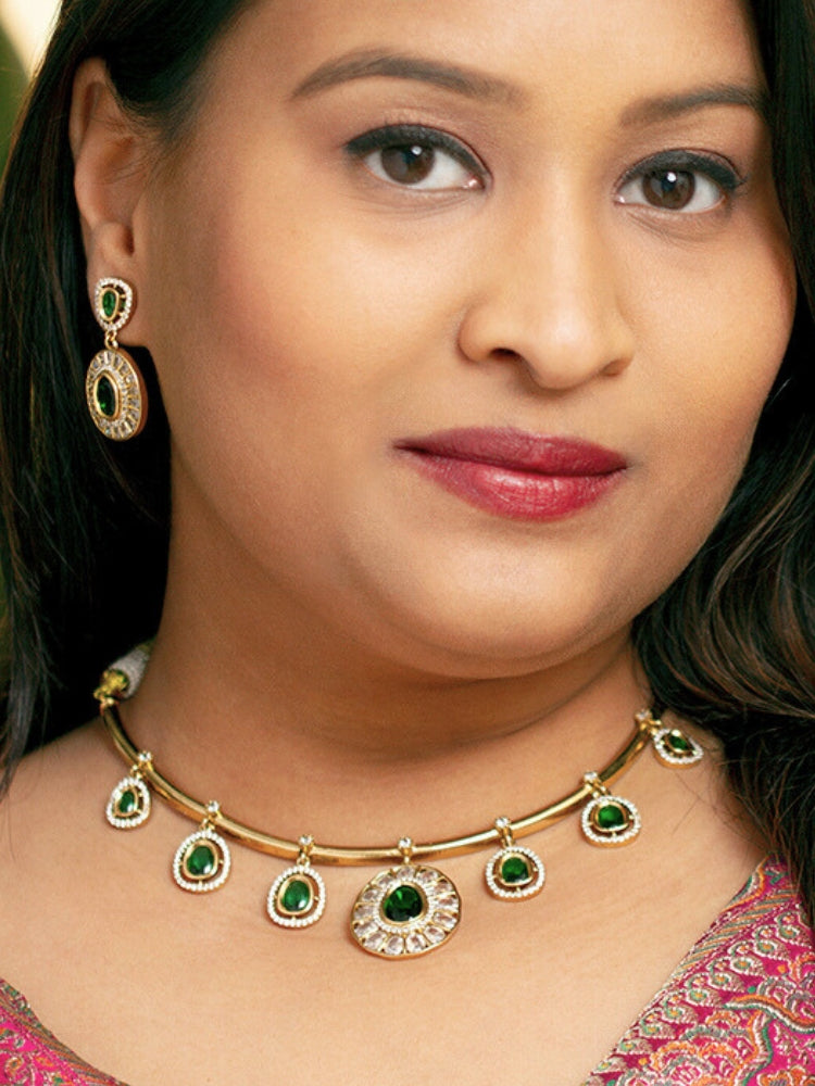 A woman with smooth skin, red lipstick, and side-parted hair wears the Tarunika Dark Green Hasli Necklace Set, featuring a vibrant White hasli necklace with Dark Green colored stone drops surrounded by american diamonds and matching earrings. The focus is on her striking jewelry.