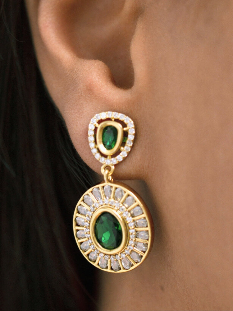 A woman with smooth skin, red lipstick, and side-parted hair wears the Tarunika Dark Green Hasli Necklace Set, featuring a vibrant White hasli necklace with Dark Green colored stone drops surrounded by american diamonds and matching earrings. The focus is on her striking jewelry.