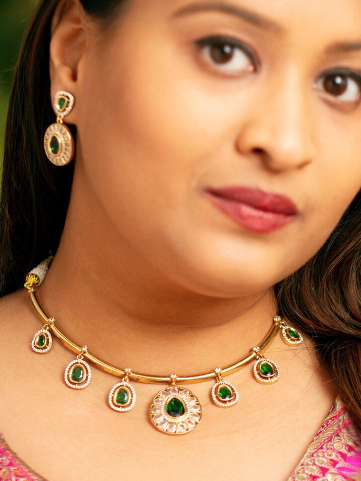 A woman with smooth skin, red lipstick, and side-parted hair wears the Tarunika Dark Green Hasli Necklace Set, featuring a vibrant White hasli necklace with Dark Green colored stone drops surrounded by american diamonds and matching earrings. The focus is on her striking jewelry.