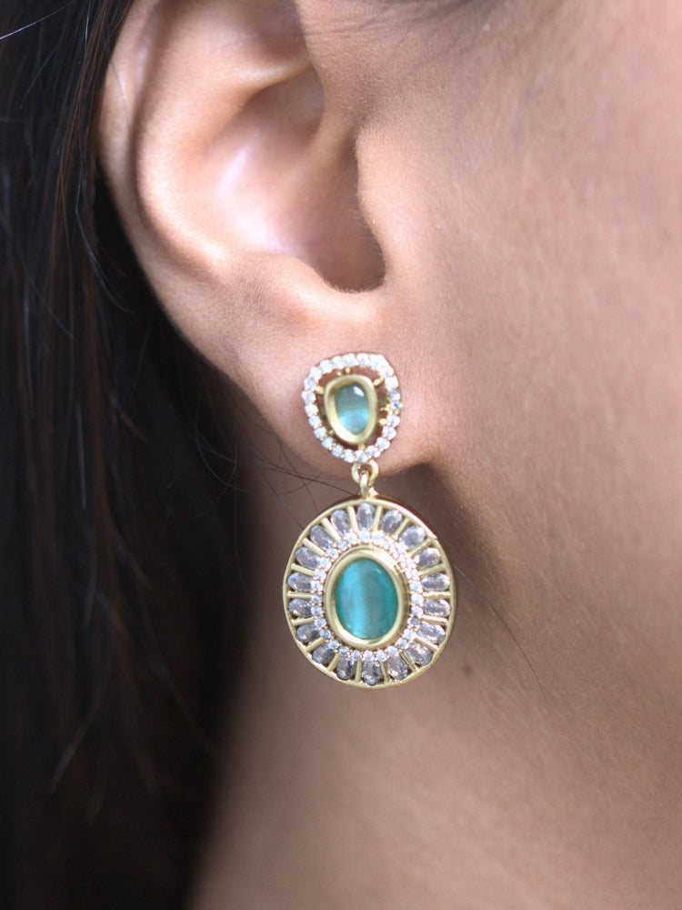 A woman with smooth skin, and side-parted hair wears the Tarunika mint green Hasli Necklace Set, featuring a vibrant purple hasli necklace with mint green colored stone drops surrounded by american diamonds and matching earrings. The focus is on her striking jewelry.