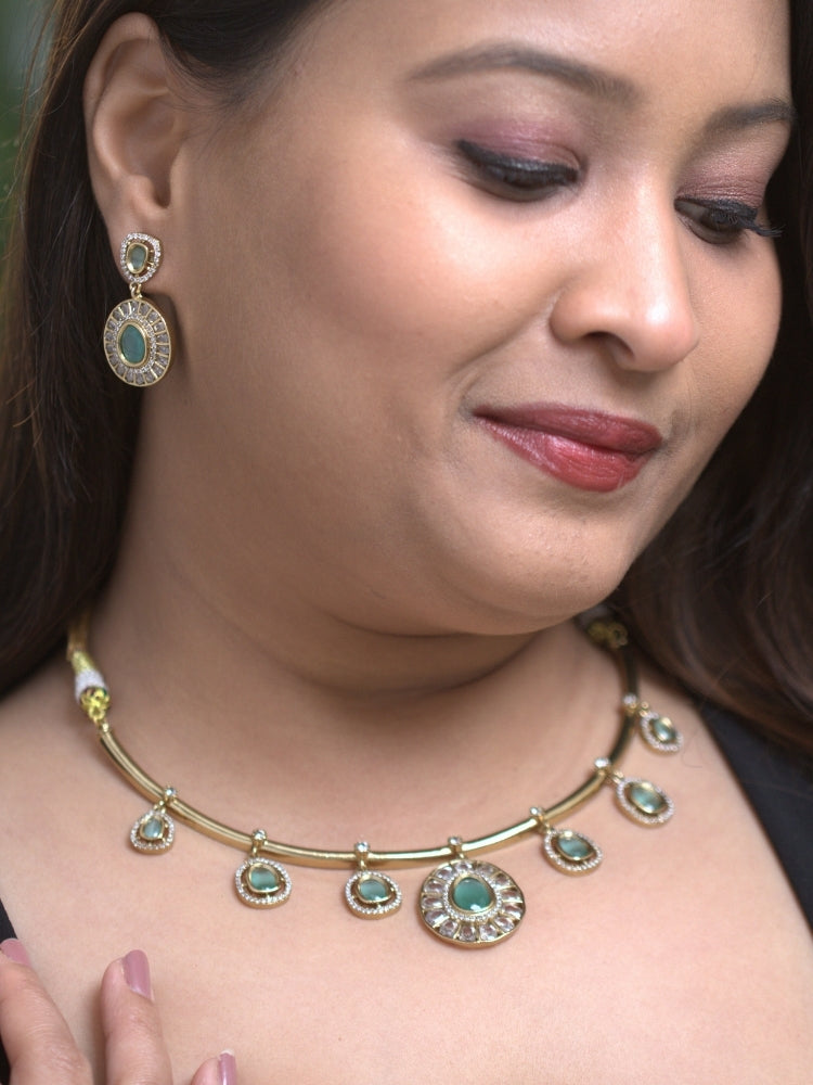 A woman with smooth skin, and side-parted hair wears the Tarunika mint green  Hasli Necklace Set, featuring a vibrant purple hasli necklace with mint green colored stone drops surrounded by american diamonds and matching earrings. The focus is on her striking jewelry.