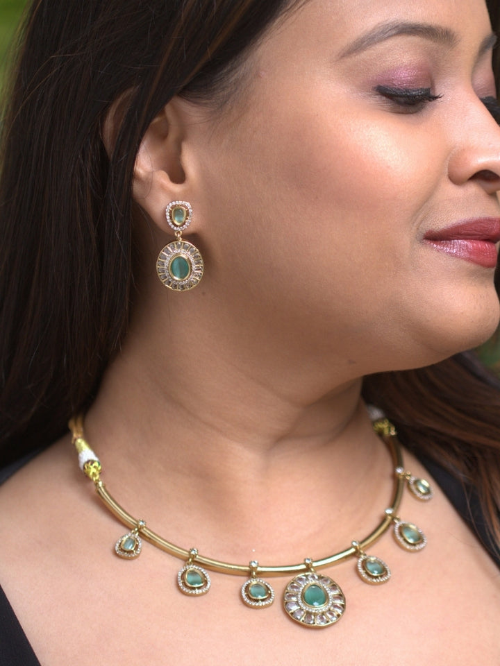A woman with smooth skin, and side-parted hair wears the Tarunika mint green Hasli Necklace Set, featuring a vibrant purple hasli necklace with mint green  colored stone drops surrounded by american diamonds and matching earrings. The focus is on her striking jewelry.