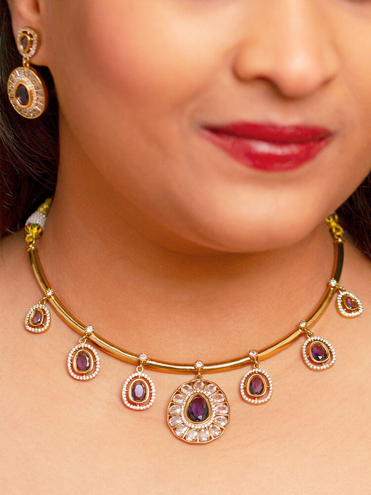 A woman with smooth skin, red lipstick, and side-parted hair wears the Tarunika Purple Hasli Necklace Set, featuring a vibrant purple hasli necklace with purple colored stone drops surrounded by american diamonds and matching earrings. The focus is on her striking jewelry.