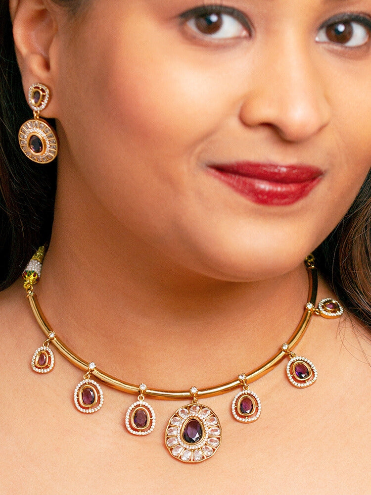 A woman with smooth skin, red lipstick, and side-parted hair wears the Tarunika Purple Hasli Necklace Set, featuring a vibrant purple hasli necklace with purple colored stone drops surrounded by american diamonds and matching earrings. The focus is on her striking jewelry.