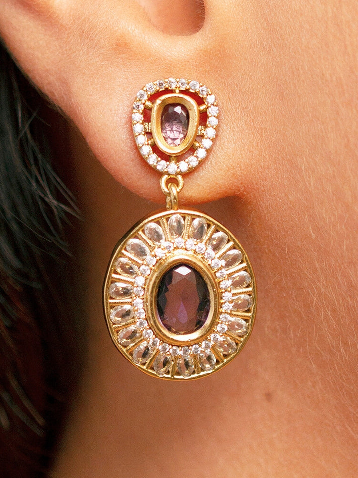 A woman with smooth skin, red lipstick, and side-parted hair wears the Tarunika Purple Hasli Necklace Set, featuring a vibrant purple hasli necklace with purple colored stone drops surrounded by american diamonds and matching earrings. The focus is on her striking jewelry.