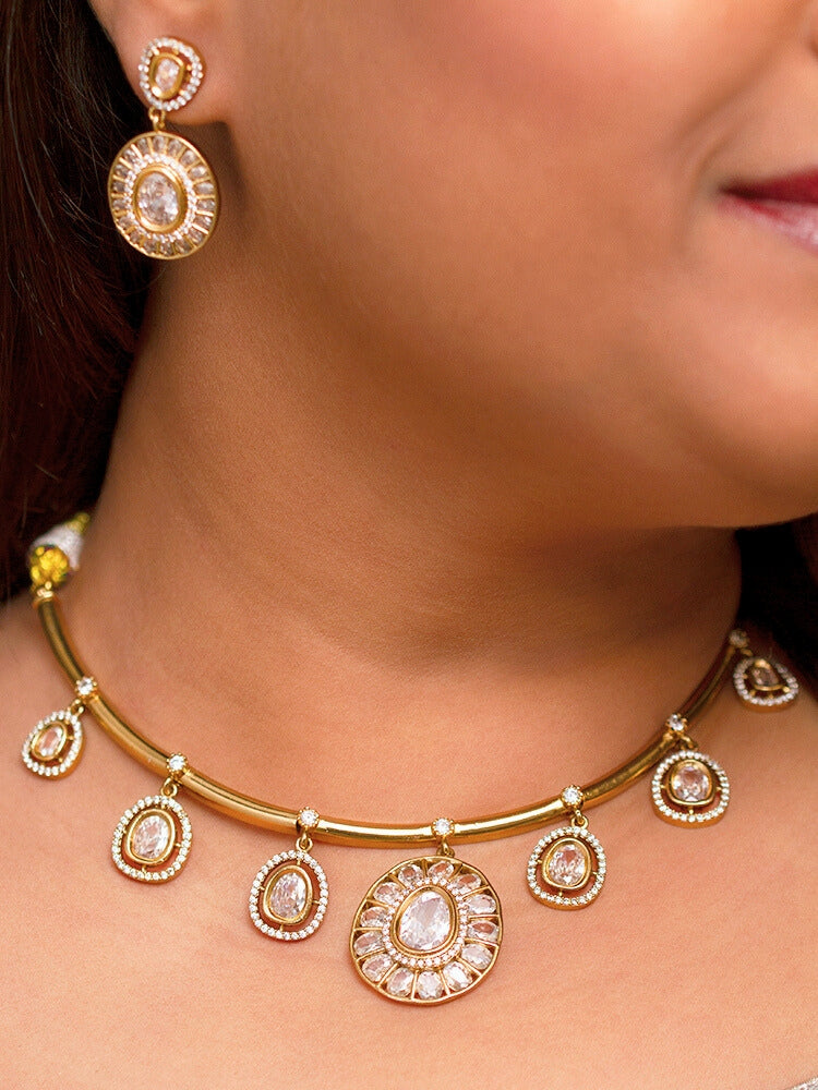 A woman with smooth skin, red lipstick, and side-parted hair wears the Tarunika White Hasli Necklace Set, featuring a vibrant White hasli necklace with White colored stone drops surrounded by american diamonds and matching earrings. The focus is on her striking jewelry.
