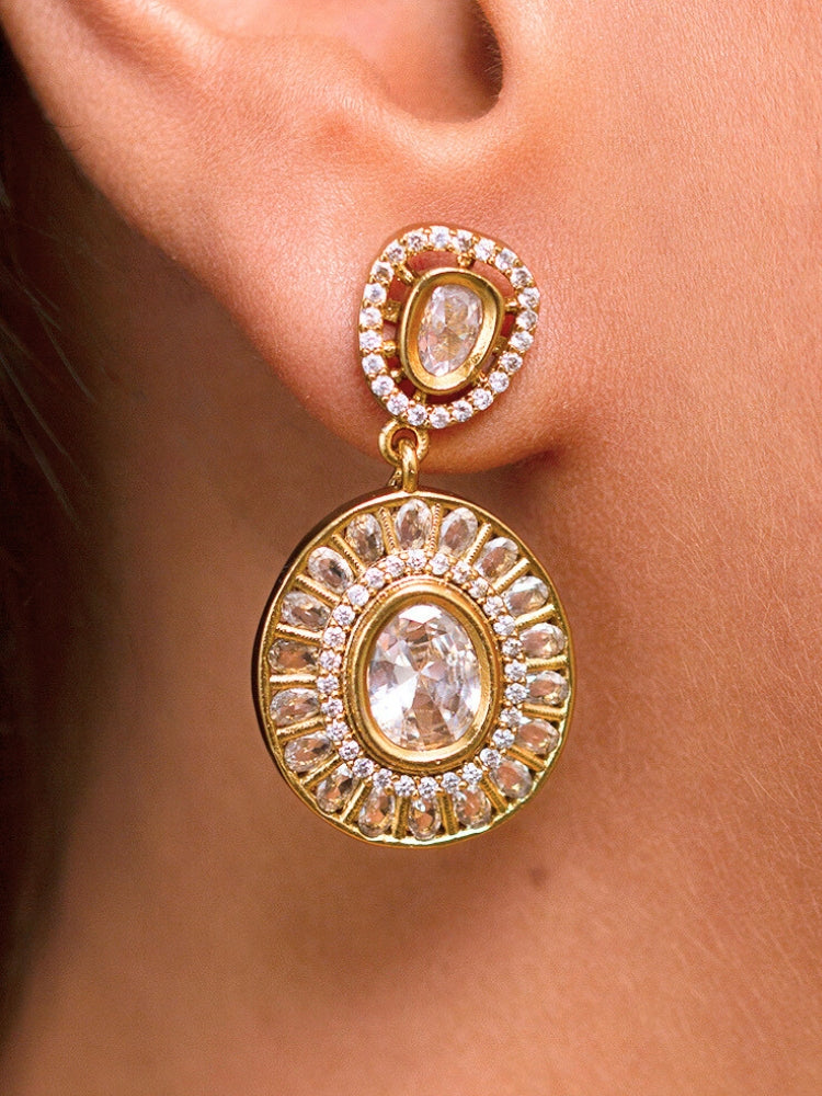 A woman with smooth skin, red lipstick, and side-parted hair wears the Tarunika White Hasli Necklace Set, featuring a vibrant White hasli necklace with White colored stone drops surrounded by american diamonds and matching earrings. The focus is on her striking jewelry.