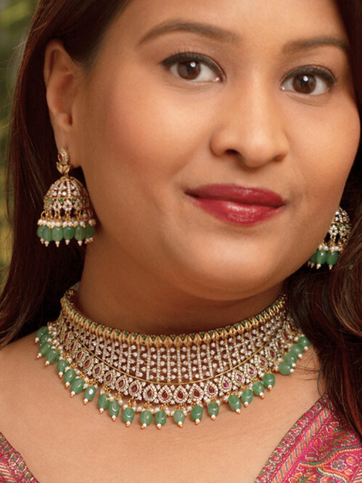 A woman wearing a maroon outfit and the Tazira Choker Necklace Set, featuring green colored stones and gold-green jhumka earrings with American diamonds, smiles softly outdoors.
