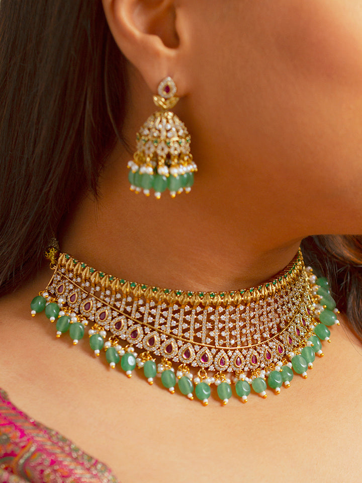 A woman wearing a maroon outfit and the Tazira Choker Necklace Set, featuring green colored stones and gold-green jhumka earrings with American diamonds, smiles softly outdoors.