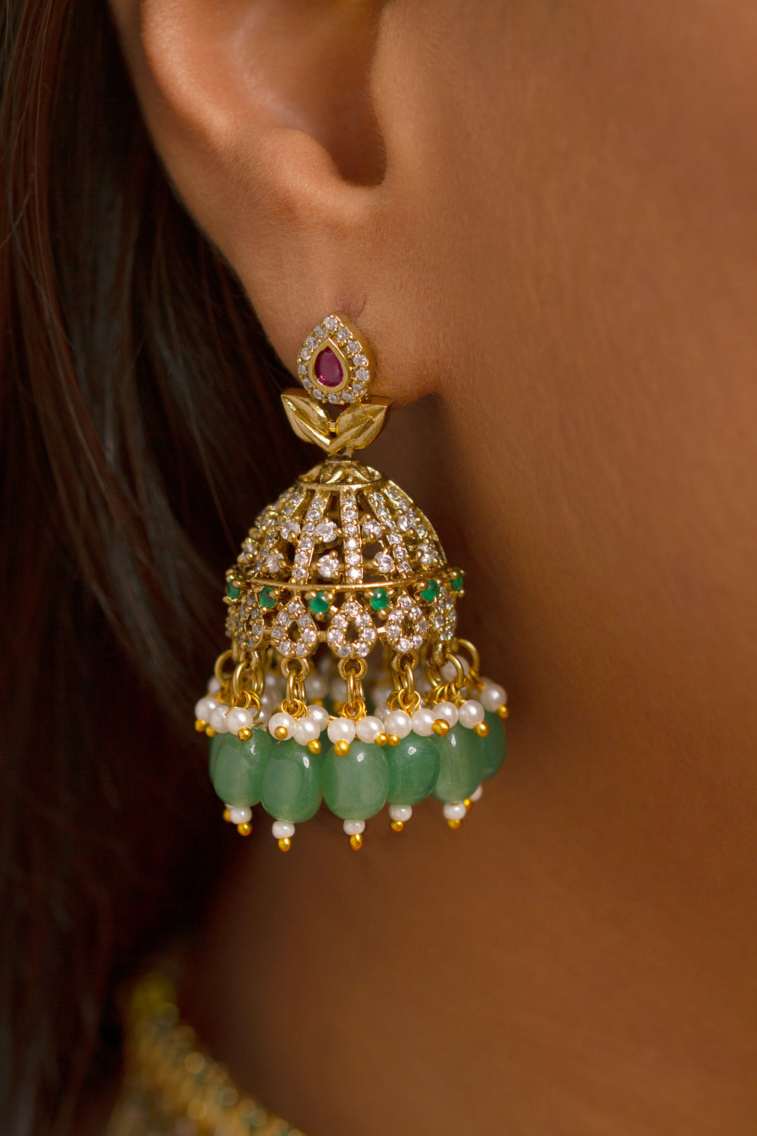 A woman wearing a maroon outfit and the Tazira Choker Necklace Set, featuring green colored stones and gold-green jhumka earrings with American diamonds, smiles softly outdoors.