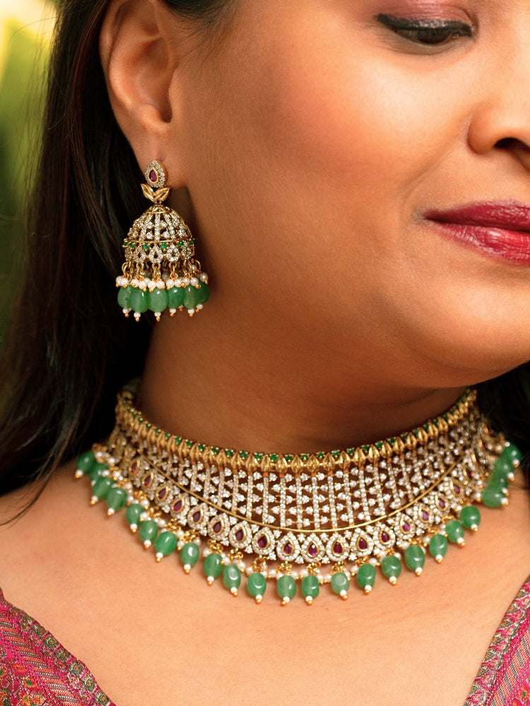 A woman wearing a maroon outfit and the Tazira Choker Necklace Set, featuring green colored stones and gold-green jhumka earrings with American diamonds, smiles softly outdoors.