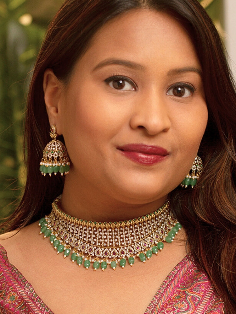 A woman wearing a maroon outfit and the Tazira Choker Necklace Set, featuring green colored stones and gold-green jhumka earrings with American diamonds, smiles softly outdoors.