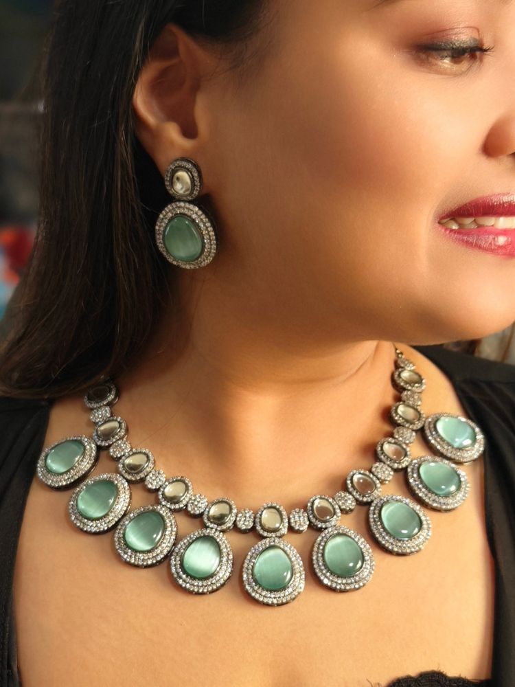 A woman with brown hair down smiles to the side, wearing the Teel Green Elizabeth Necklace Set featuring a statement Victorian-style necklace and matching earrings with large colorful stones, paired with a sparkly dark top.