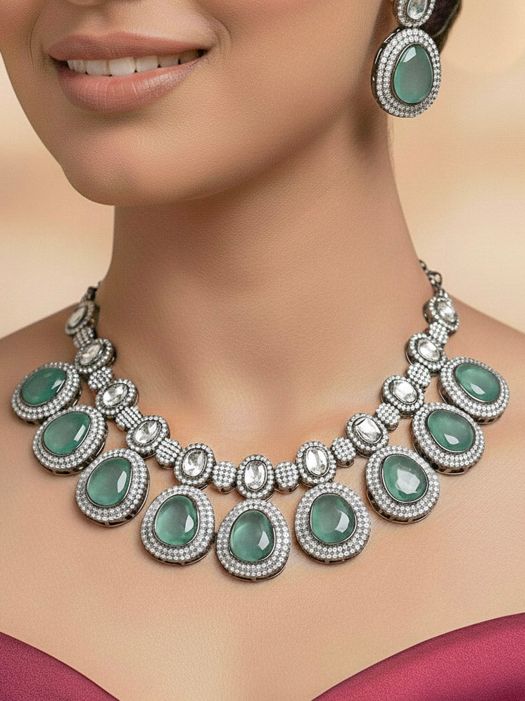 A woman with brown hair down smiles to the side, wearing the Teel Green Elizabeth Necklace Set featuring a statement Victorian-style necklace and matching earrings with large colorful stones, paired with a sparkly dark top.