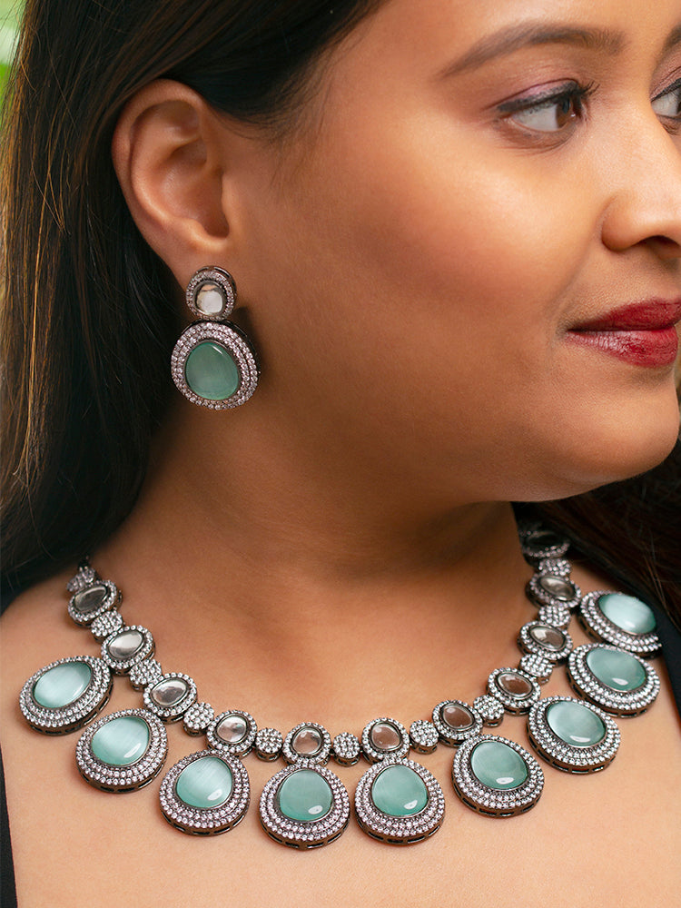 A woman with brown hair down smiles to the side, wearing the Teel Green Elizabeth Necklace Set featuring a statement Victorian-style necklace and matching earrings with large colorful stones, paired with a sparkly dark top.