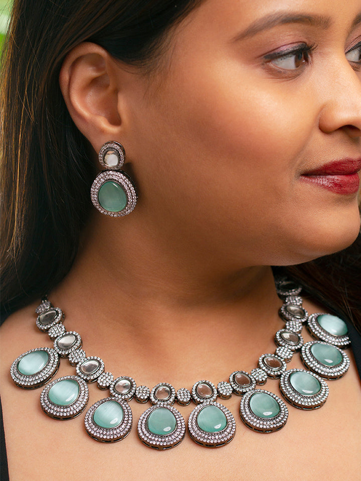 A woman with brown hair down smiles to the side, wearing the Teel Green Elizabeth Necklace Set featuring a statement Victorian-style necklace and matching earrings with large colorful stones, paired with a sparkly dark top.