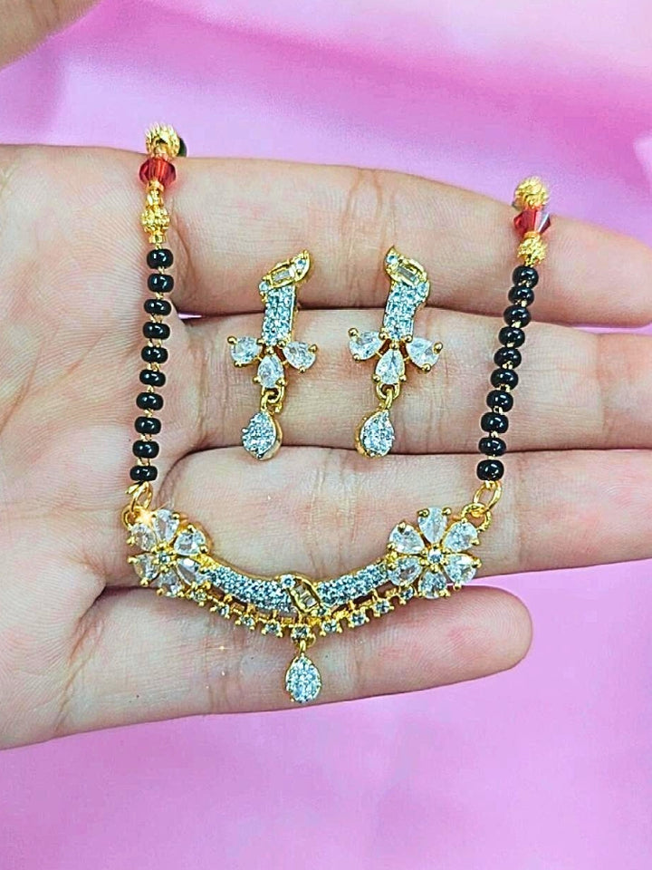 A hand holds the Tina Mangalsutra—a gold necklace with black mangalsutra beads and matching earrings, pendant featuring sparkling American Diamonds—set against a soft pink background, perfect for your wedding ceremony dress. Diamond mangalsutra. Modern mangalsutra. 