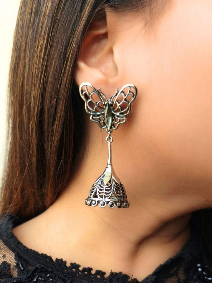A close-up of a womans ear adorned with Titli Earrings—large, shiny German Silver earrings featuring a butterfly design on top and a jhumka-style bell below. Ideal for wedding ceremonies.