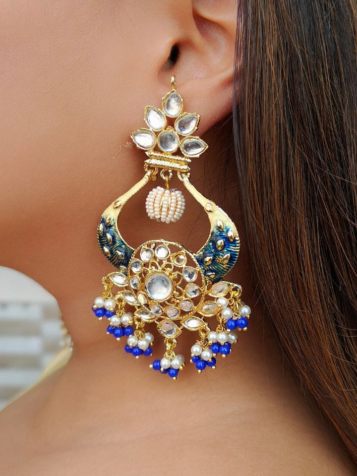 A close-up of a womans ear wearing Udaya Earrings—ornate gold Kundan design with meenakari work,  American diamonds, small pearls, blue beads, and floral details—shown against her straight brown hair.