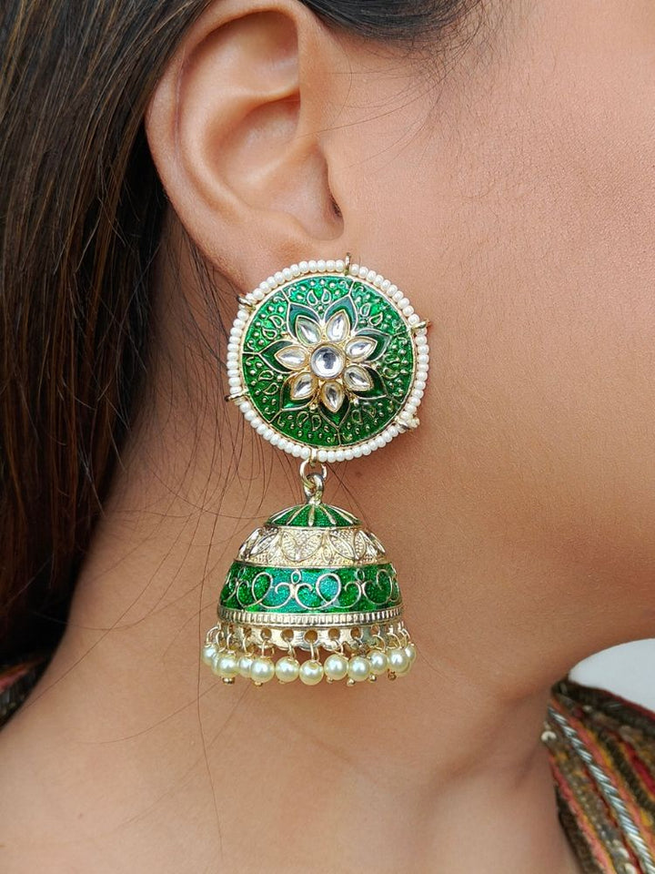 A close-up of a womans ear wearing the Upasna Earrings, featuring large green and gold circular jhumkas with pearl accents, floral patterns, and exquisite Meena work.