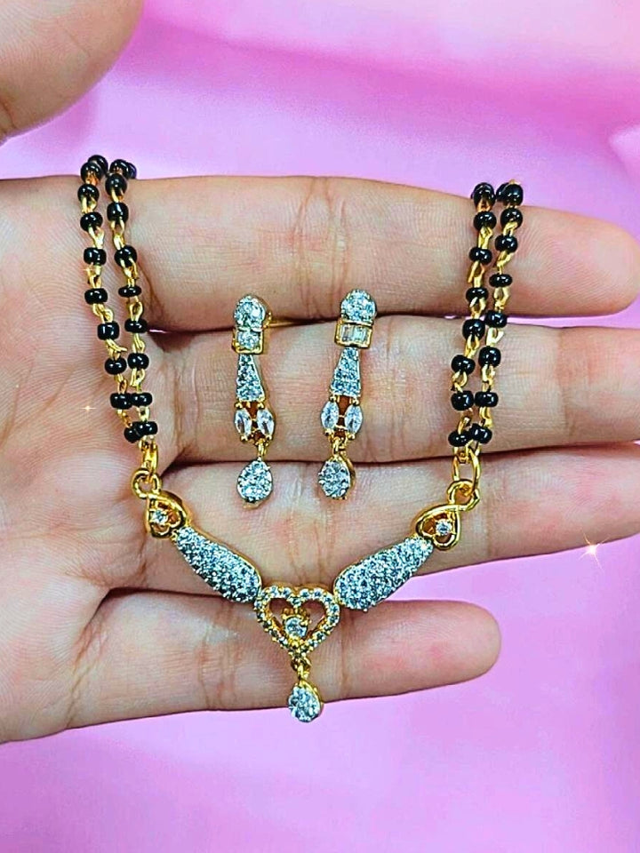 A hand holds the Urvashi Mangalsutra—a gold necklace with black mangalsutra beads and matching earrings, pendant featuring sparkling American Diamonds—set against a soft pink background, perfect for your wedding ceremony dress. Diamond mangalsutra. Modern mangalsutra. 