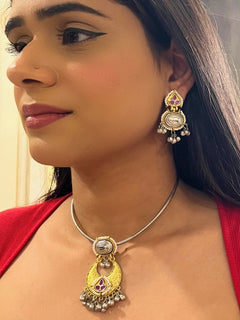 A woman wears the Usha Pink Necklace Set, featuring colored stones and matching earrings. The brass pendant oxidized necklace pops against her red top, with just her lower face, neck, and upper chest in view