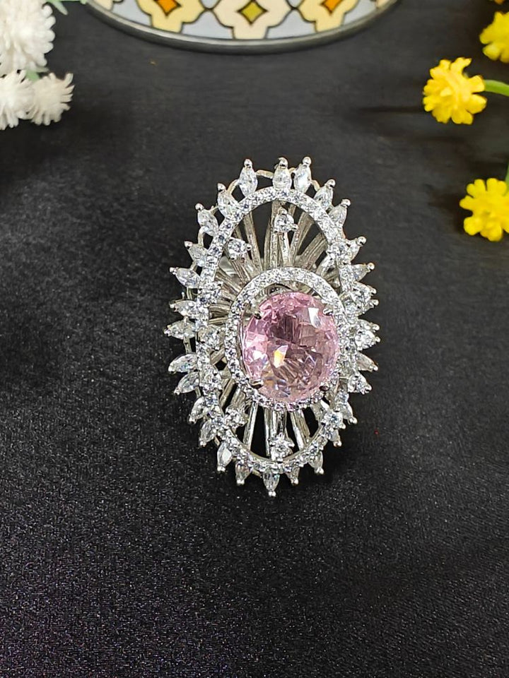 The Valentina Pink Center Stone Ring features an ornate large round pink gem at its center, encircled by sparkling American Diamonds and set on an adjustable band, beautifully displayed on a black surface with yellow flowers. American Diamond Cocktail Ring, American Diamond Ring. 