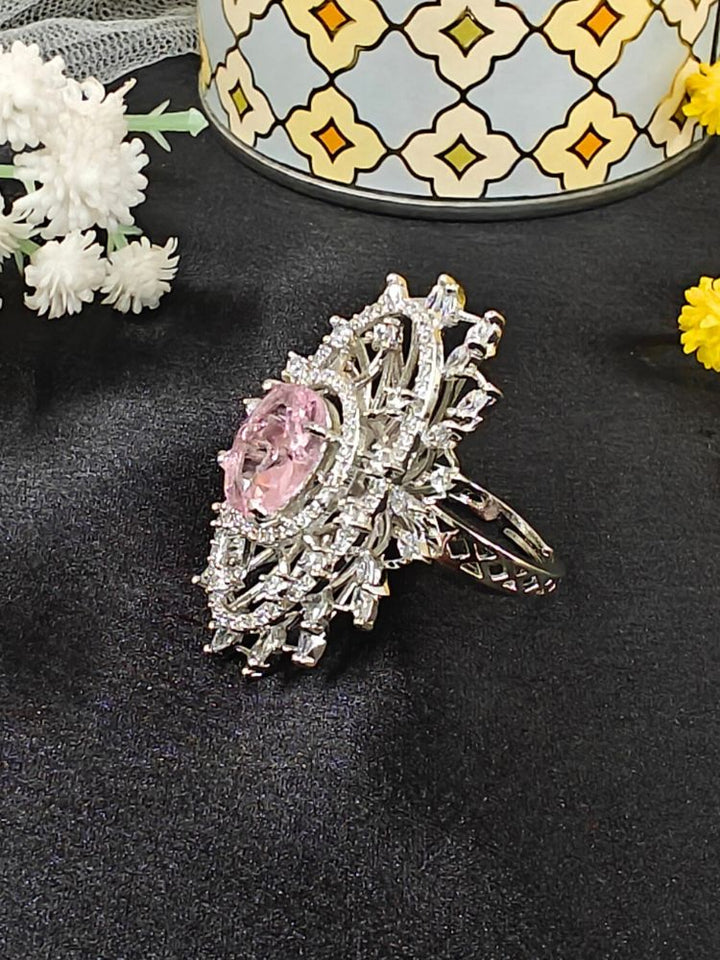 The Valentina Pink Center Stone Ring features an ornate large round pink gem at its center, encircled by sparkling American Diamonds and set on an adjustable band, beautifully displayed on a black surface with yellow flowers. American Diamond Cocktail Ring, American Diamond Ring. 