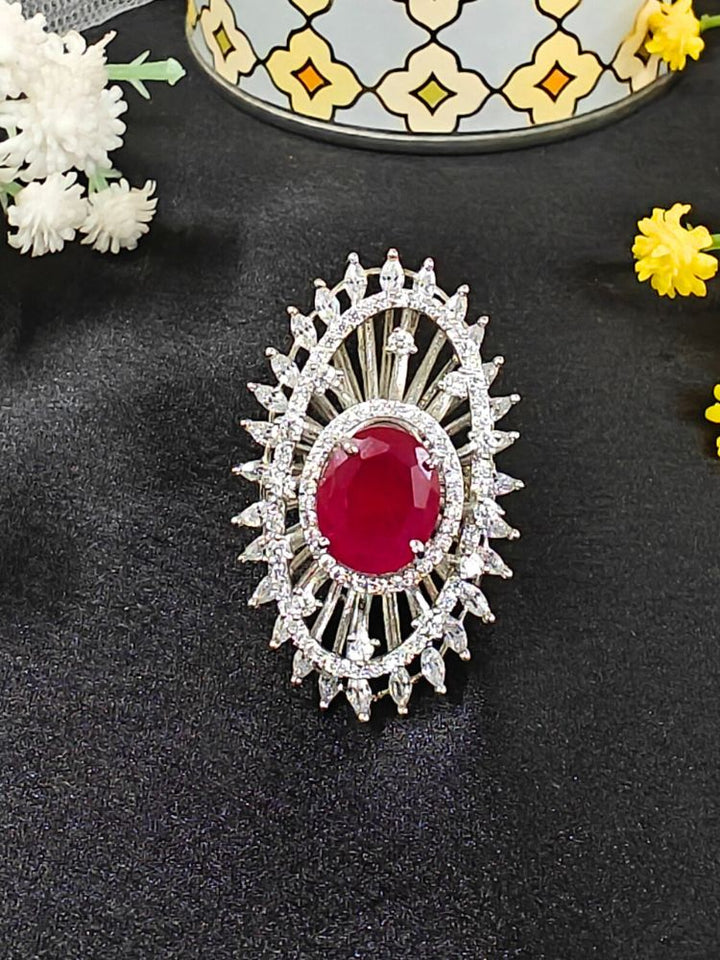 The Valentina Ruby Red Center Stone Ring features an ornate large round Ruby Red gem at its center, encircled by sparkling American Diamonds and set on an adjustable band, beautifully displayed on a black surface with yellow flowers. American Diamond Cocktail Ring, American Diamond Ring. 