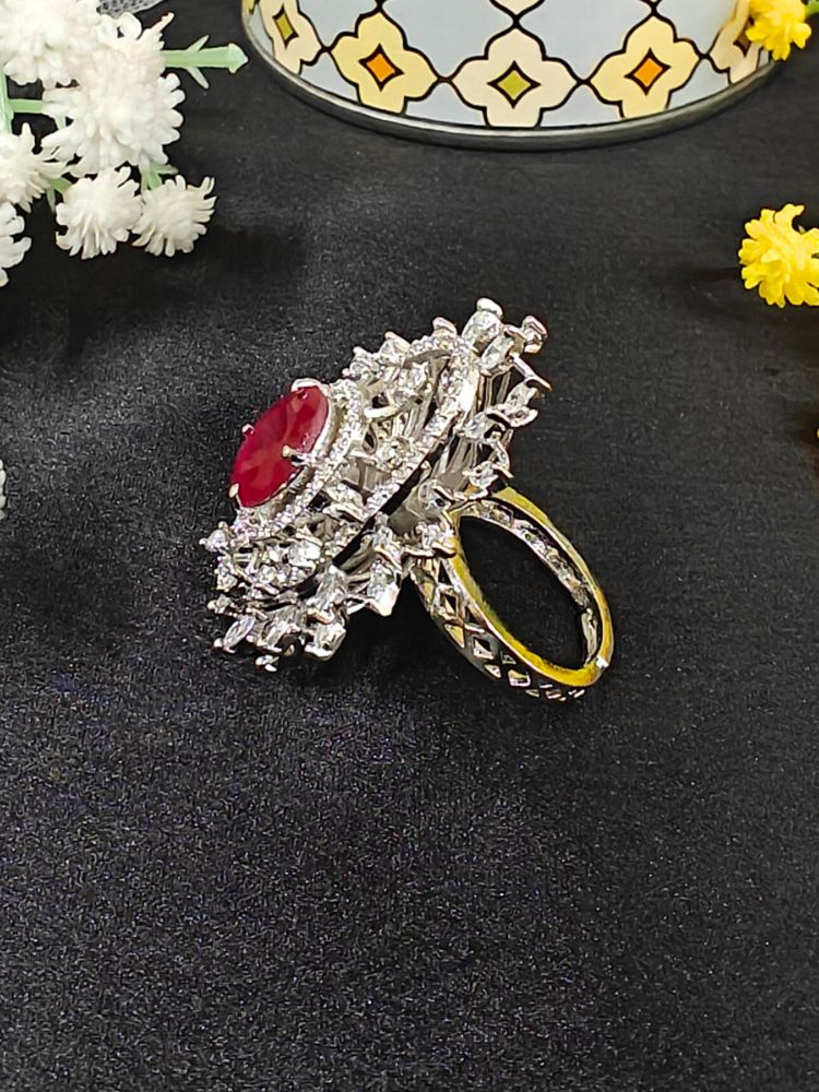 The Valentina Ruby Red Center Stone Ring features an ornate large round Ruby Red gem at its center, encircled by sparkling American Diamonds and set on an adjustable band, beautifully displayed on a black surface with yellow flowers. American Diamond Cocktail Ring, American Diamond Ring. 