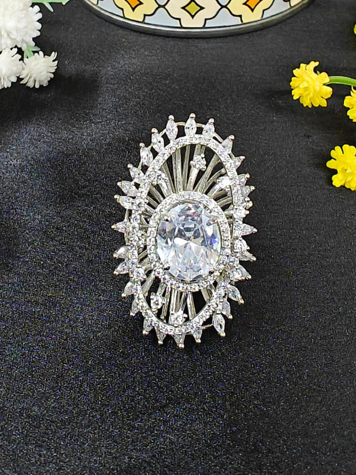 The Valentina White Center Stone Ring features an ornate large round White gem at its center, encircled by sparkling American Diamonds and set on an adjustable band, beautifully displayed on a black surface with yellow flowers. American Diamond Cocktail Ring, American Diamond Ring. 