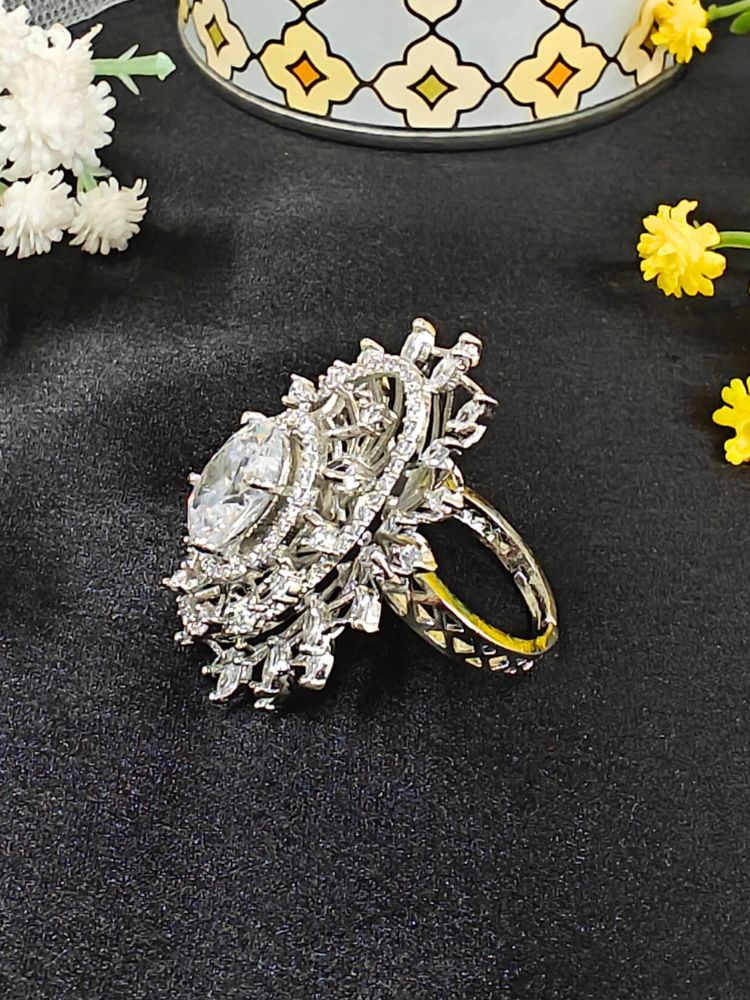 The Valentina White Center Stone Ring features an ornate large round White gem at its center, encircled by sparkling American Diamonds and set on an adjustable band, beautifully displayed on a black surface with yellow flowers. American Diamond Cocktail Ring, American Diamond Ring. 