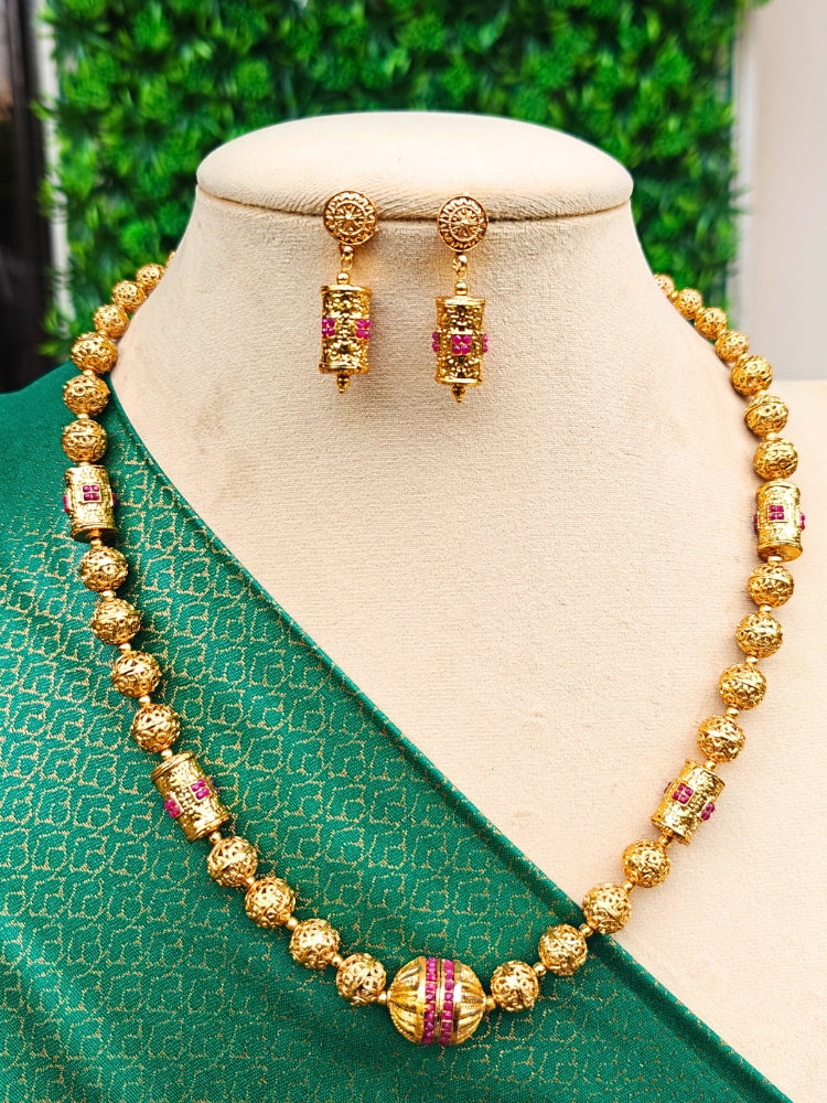 The Varsha Necklace Set, featuring gold matar mala, with  ruby red stones accentuating the golf matar mala, along with matching drop earrings, is displayed on a mannequin in a green patterned blouse against a leafy backdrop—an elegant choice for wedding jewelry.