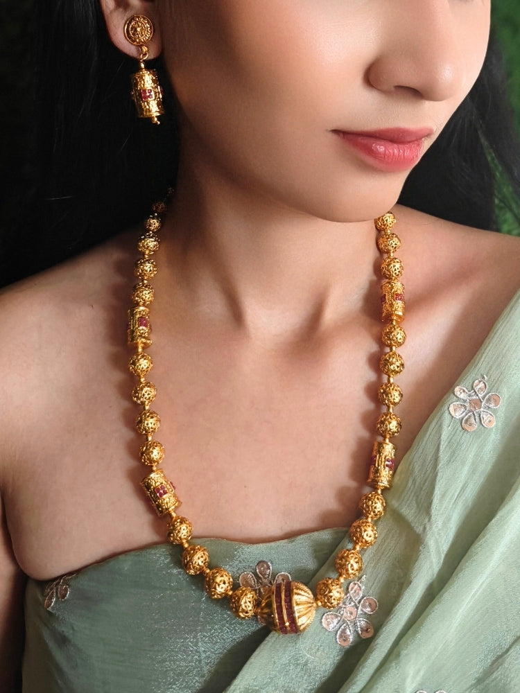 Close-up of a woman wearing an ornate traditional (Bright Gold Polish) Long Jewellery Set with Earrings with Ruby Red Colored Stones and Filigree Work. Saree look completed with an elegant Colored Stone artificial jewelry piece. Woman wearing trending Colored Stone artificial jewelry for wedding season in India.