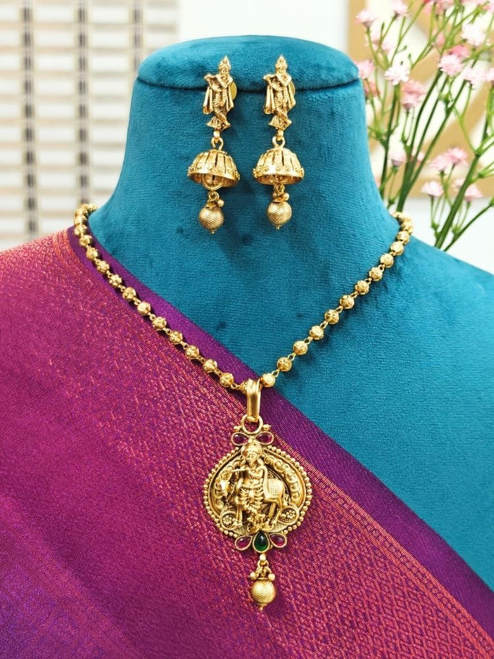 A mannequin draped in a purple and pink saree showcases the Vasudha Pendant Set—a gold deity-design necklace with matching dangling earrings—against a softly blurred, flower-filled background.