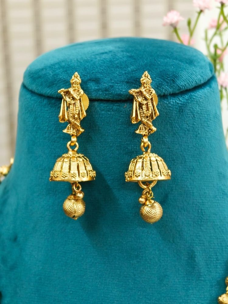 A mannequin draped in a purple and pink saree showcases the Vasudha Pendant Set—a gold deity-design necklace with matching dangling earrings—against a softly blurred, flower-filled background.