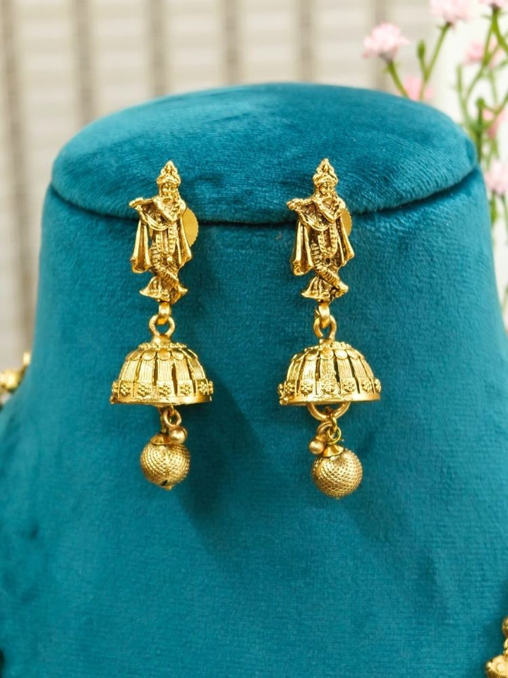 A mannequin draped in a purple and pink saree showcases the Vasudha Pendant Set—a gold deity-design necklace with matching dangling earrings—against a softly blurred, flower-filled background.