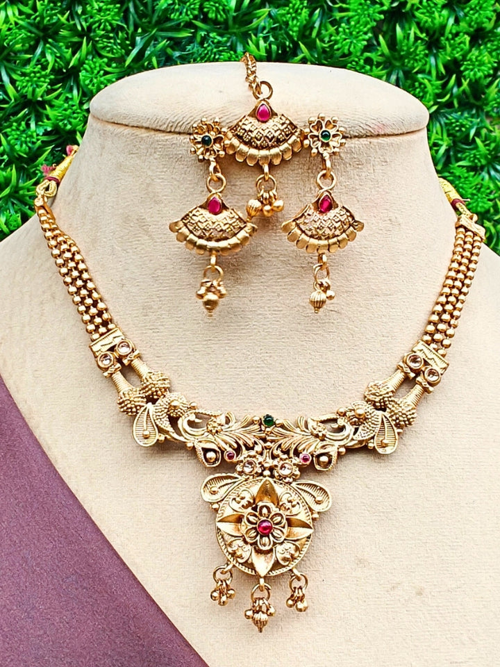 The Vedavati Necklace Set is a gold-plated South Indian necklace with an intricate floral pendant, red and green stones, and matching jhumka earrings—perfect for weddings and special occasions. Displayed on a beige jewelry bust.