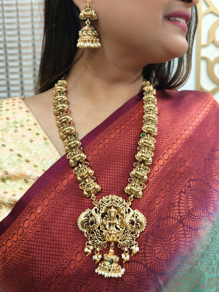 A woman in a maroon saree with gold embroidery and a cream blouse, adorned with traditional South-Indian jewelry, including the Vibhuti Temple Necklace Set featuring an ornate Laxmi necklace and matching temple jhumka earrings.